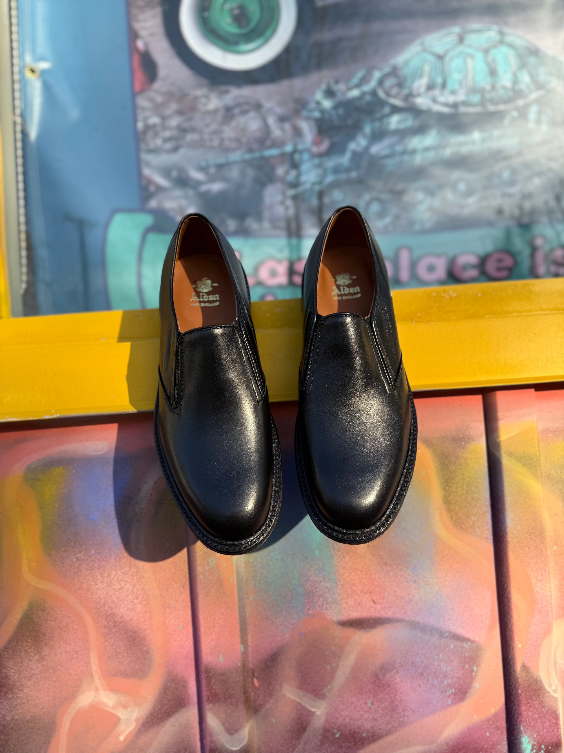 A pair of ALDEN SHOES D2406L "Double Gore" black calfskin slip-on shoes with a rubber sole rests elegantly on a yellow surface.