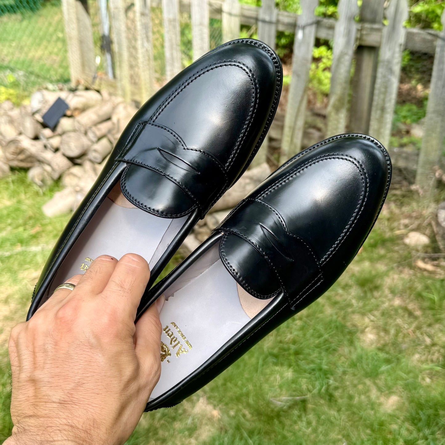 A hand holding the Alden Madison 96949 - Copley Penny Loafer in Black Shell Cordovan, featuring a classic 270 Flat Welt, outdoors against a backdrop of grass, rocks, and a wooden fence.