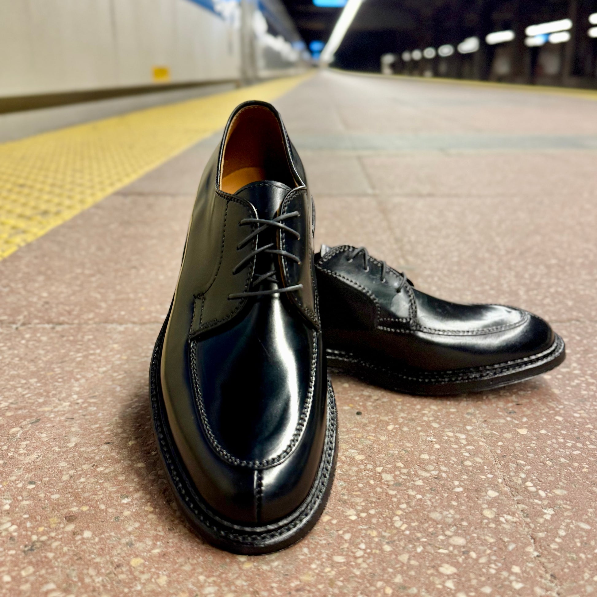 A pair of ALDEN SHOES D4604C Moc Toe “Borough” Blucher in Black Shell sits on a tiled subway platform, with yellow tactile paving and blurred lights in the background.
