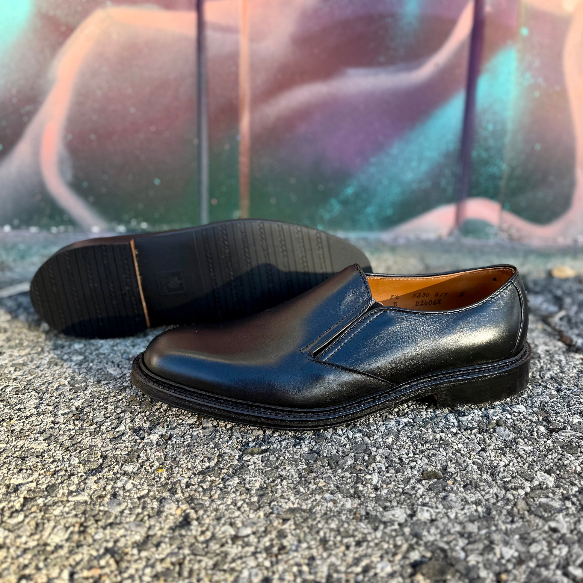 A pair of ALDEN SHOES D2406L "Double Gore" black calfskin slip-on dress shoes with rubber soles is displayed outdoors on textured ground, with one shoe upright and the other on its side before a blurred, colorful mural.