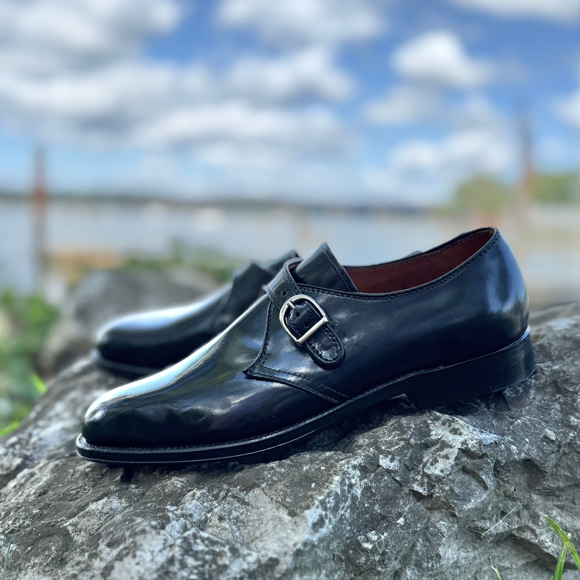 Alden Madison’s 1655 - Monk Strap Black in Shell Cordovan, featuring silver buckles and a single oak leather sole on the Aberdeen Last, sits atop a gray rock with blue sky, clouds, water, and greenery in the blurred background.