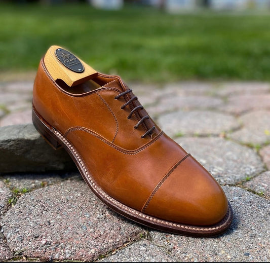 The Alden Madison 9062 - Straight Tip Bal in Burnished Tan Calfskin with a wooden shoe tree is displayed outdoors on stone pavement, its refined Single Oak Leather Sole standing out against the blurred grass background.