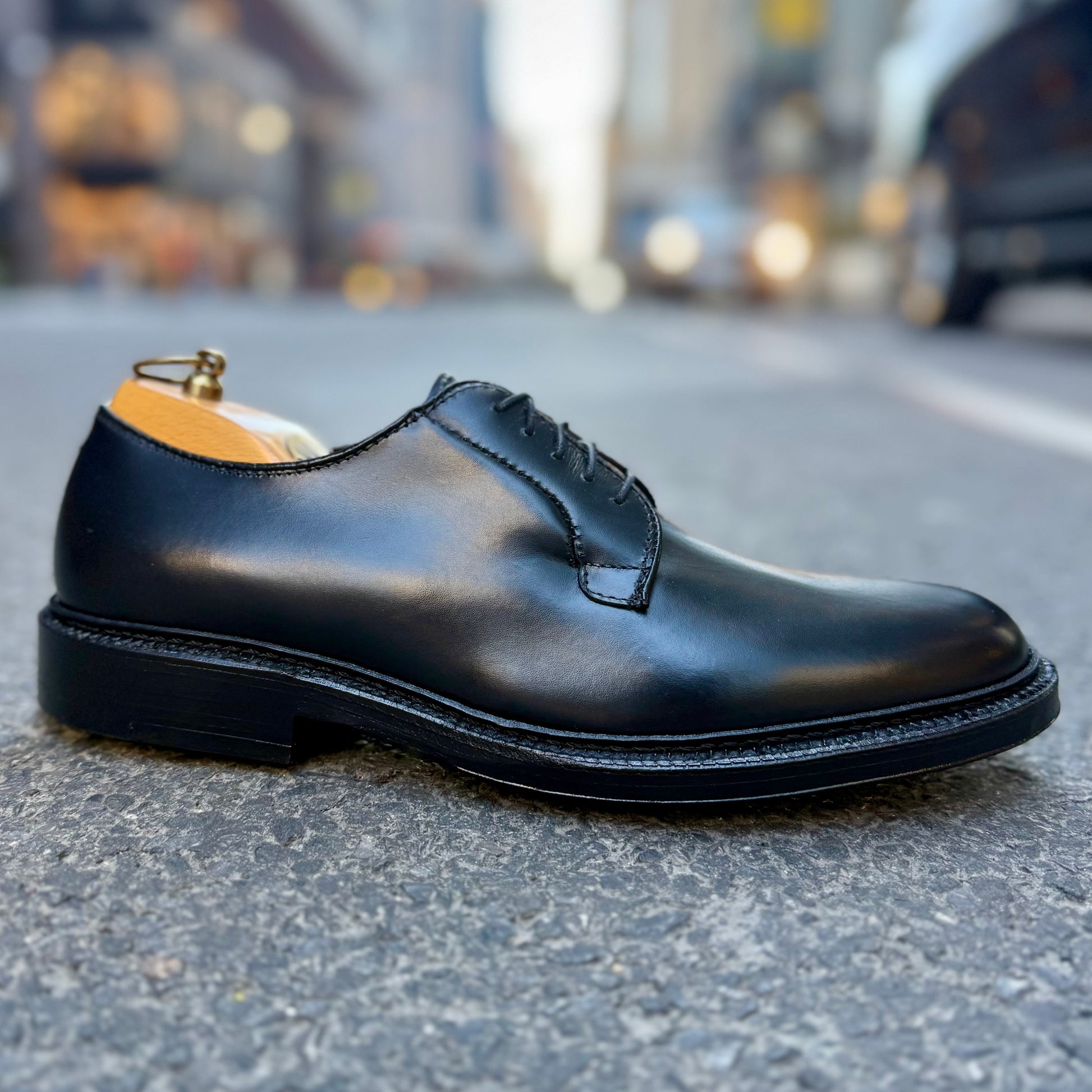 The ALDEN SHOES 9909 - Black Calfskin Plain Toe Blucher with laces is displayed on a city street, with blurred cars and buildings in the background.