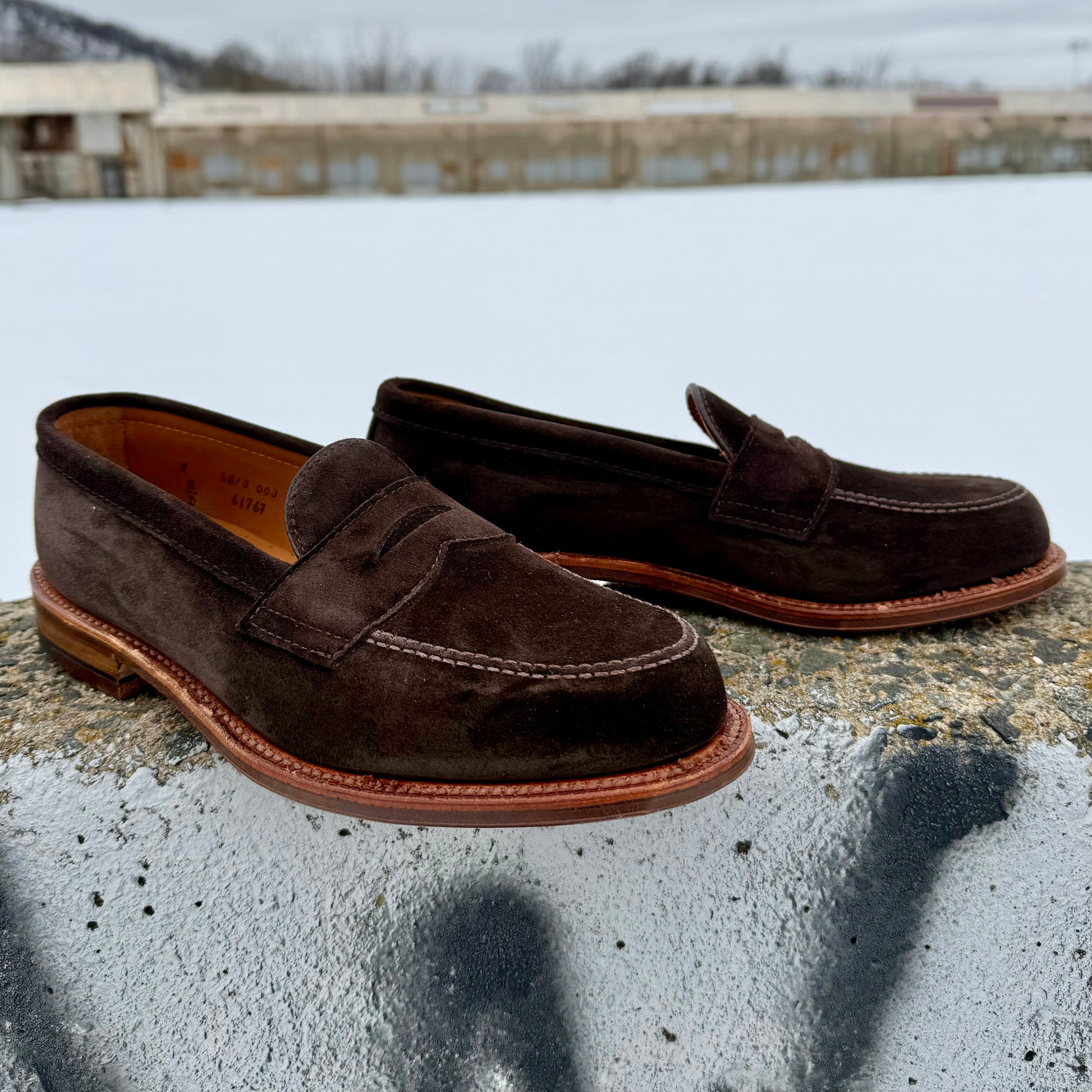 The Alden Madison 6176F Penny Loafer in Dark Chocolate Suede, with a flex oiled leather sole, sits on concrete outdoors, set against a snowy landscape and industrial buildings.