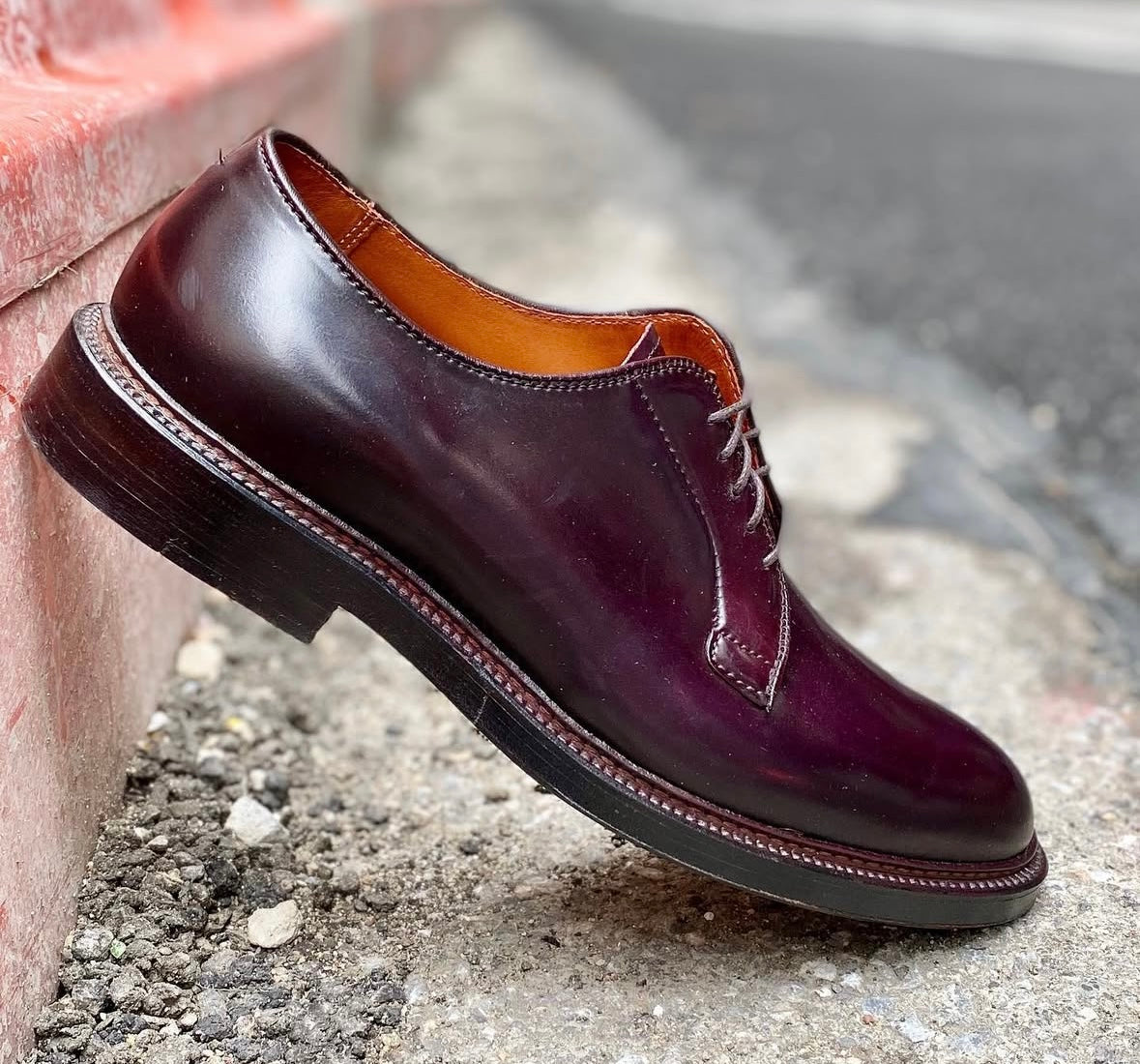 A single Alden Madison 990 - Plain Toe Blucher in Color 8 Shell Cordovan, featuring brown laces and a Double Oak Leather Sole on the Barrie Last, is propped sideways against a red curb on rough gray pavement outdoors.