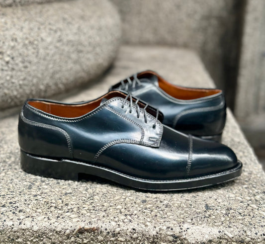 The Alden Madison 2161 - Cap Toe Blucher in Black Shell Cordovan features a sleek cap toe and lace-up design, crafted on the Aberdeen Last, and is shown on a textured stone surface.