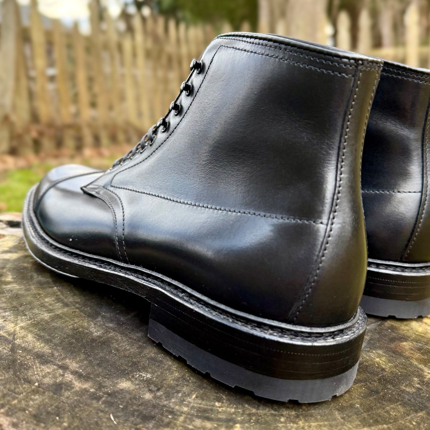 A close-up of Alden Madison’s 40797HC Black Cap Toe Boot with rugged commando sole, shown outdoors on a wooden surface with a blurred wooden fence in the background.