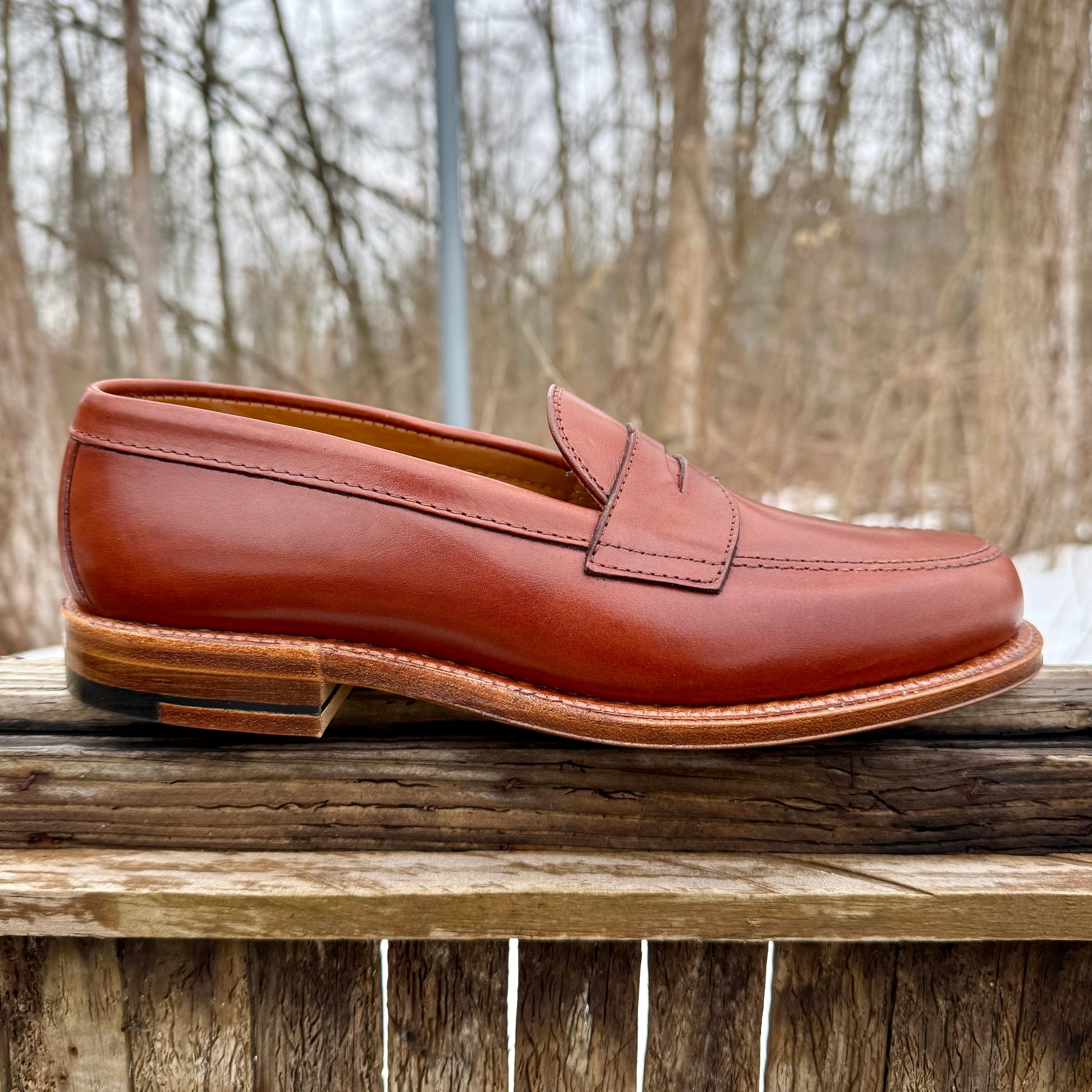 The Alden Madison 9823 - Penny Loafer in Tan Michigan Calf, featuring stitching details and an Oak Leather Sole, is shown outdoors on a wooden surface with bare trees and an overcast sky in the background.