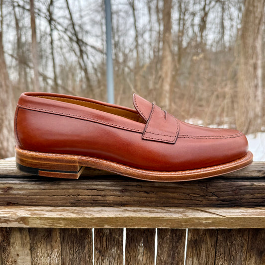 The Alden Madison 9823 - Penny Loafer in Tan Michigan Calf, featuring stitching details and an Oak Leather Sole, is shown outdoors on a wooden surface with bare trees and an overcast sky in the background.