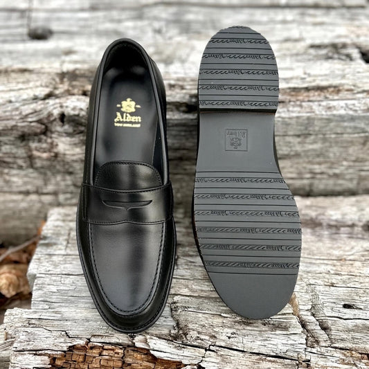 The ALDEN SHOES D5912L penny loafer in black lady calf is shown on weathered wood, with one shoe displaying the top and the other highlighting its durable rubber sole with detailed tread patterns and branding.