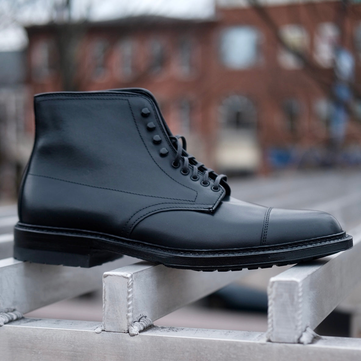 The Alden Madison 40797HC Black Cap Toe Boot with Commando Sole is shown outdoors on a metal surface, with a blurred backdrop of brick buildings and trees.