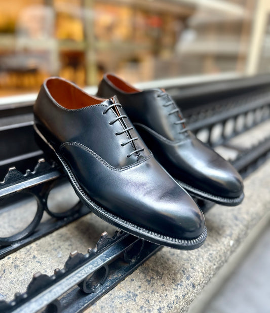 The Alden Madison 932 - Plain Toe Bal in Black Calfskin, featuring the elegant Aberdeen Last and a classic leather sole, is showcased outdoors on a stone surface, leaning against a decorative metal railing.