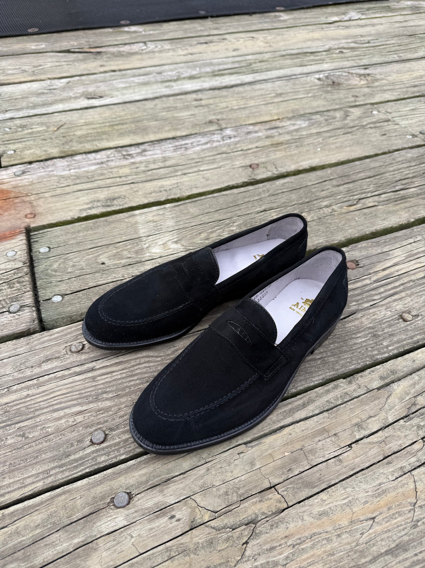 The Alden Madison D2229 “Stuyvesant” Penny in black suede sits side by side on a weathered wooden deck, white interiors and leather soles subtly contrasting with the rustic boards.