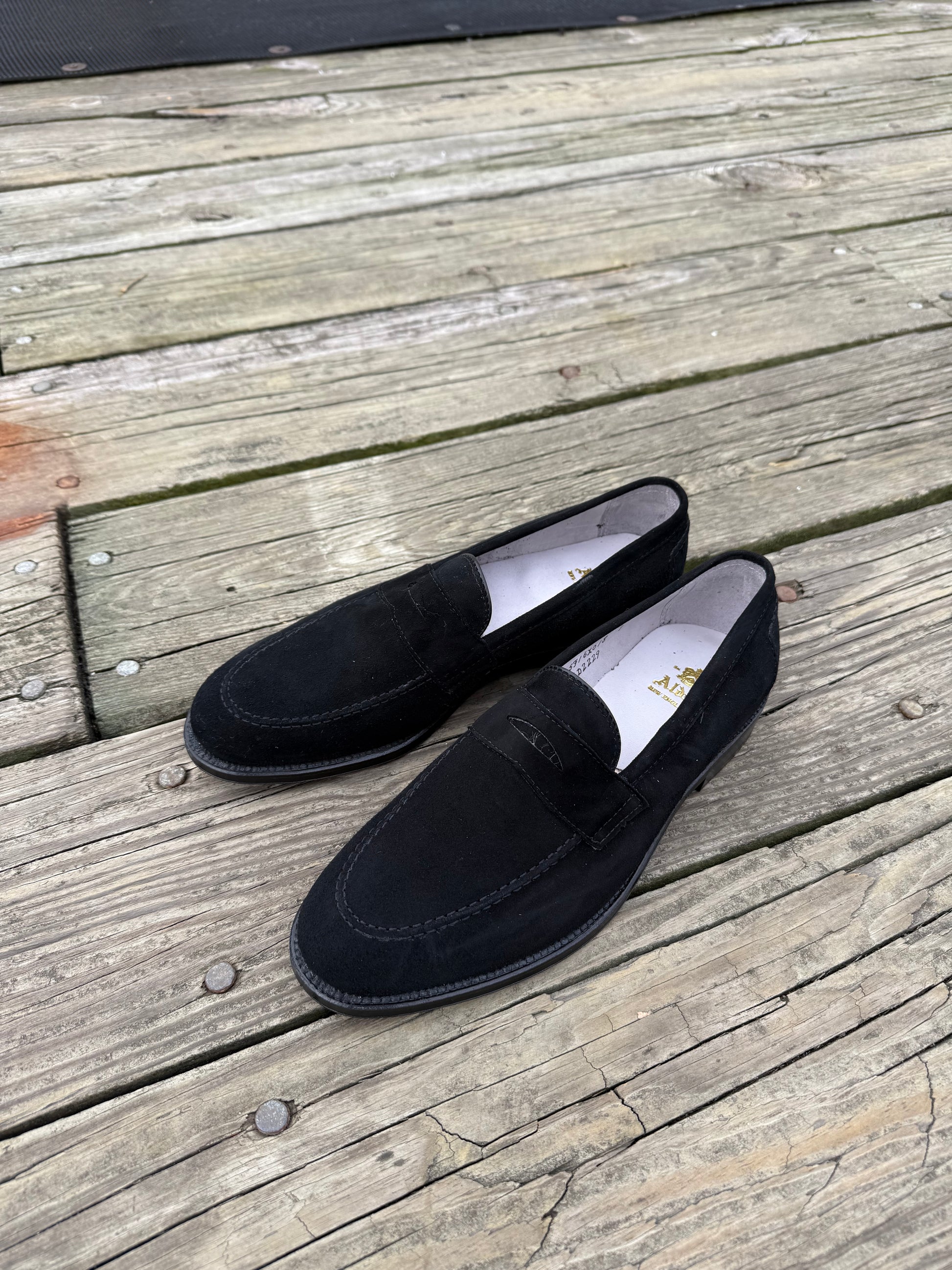The Alden Madison D2229 “Stuyvesant” Penny in black suede sits side by side on a weathered wooden deck, white interiors and leather soles subtly contrasting with the rustic boards.