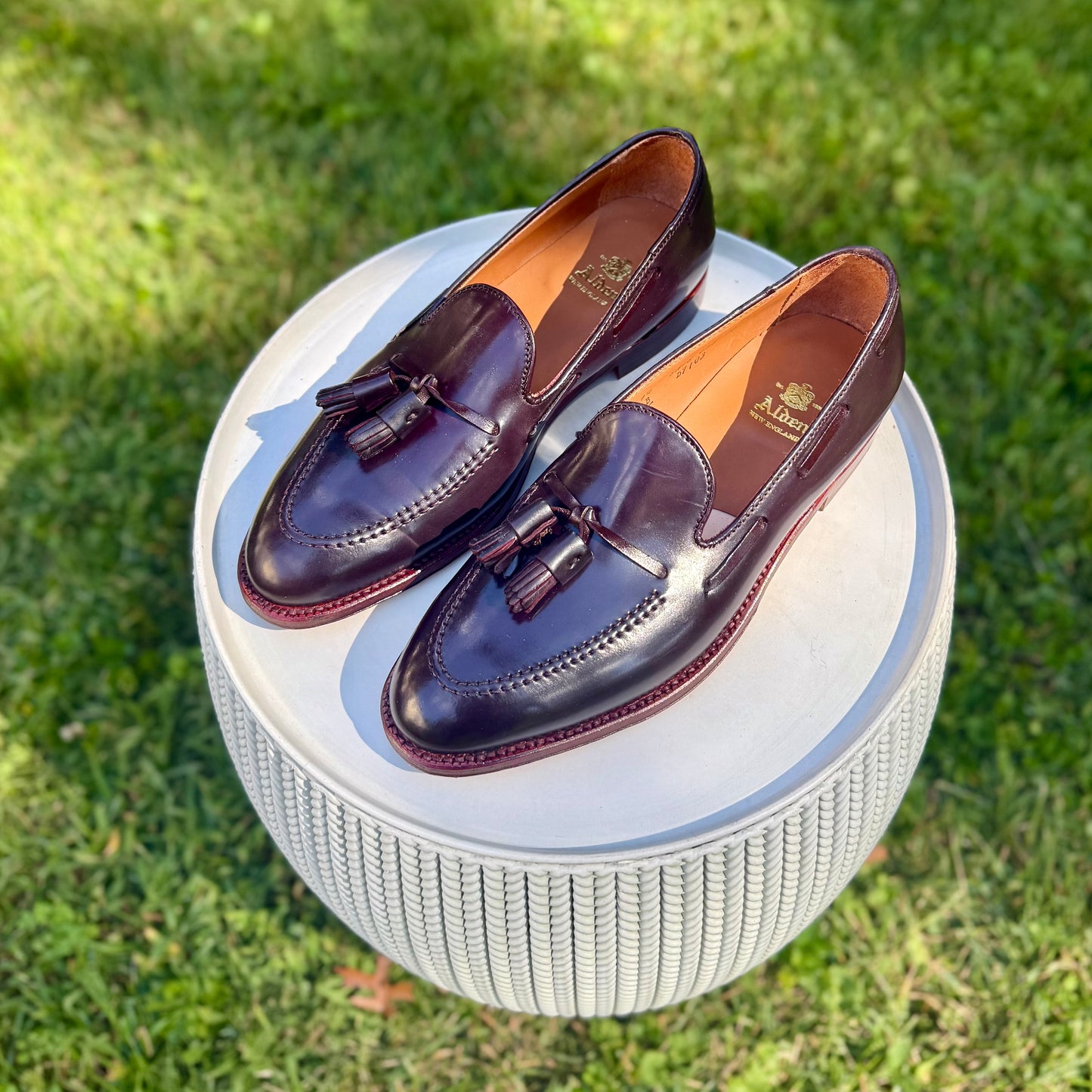 The Alden Madison D7103 - Antique Tassel Loafer in Color 8 Shell Cordovan is displayed on a round, textured white stool outdoors on green grass.