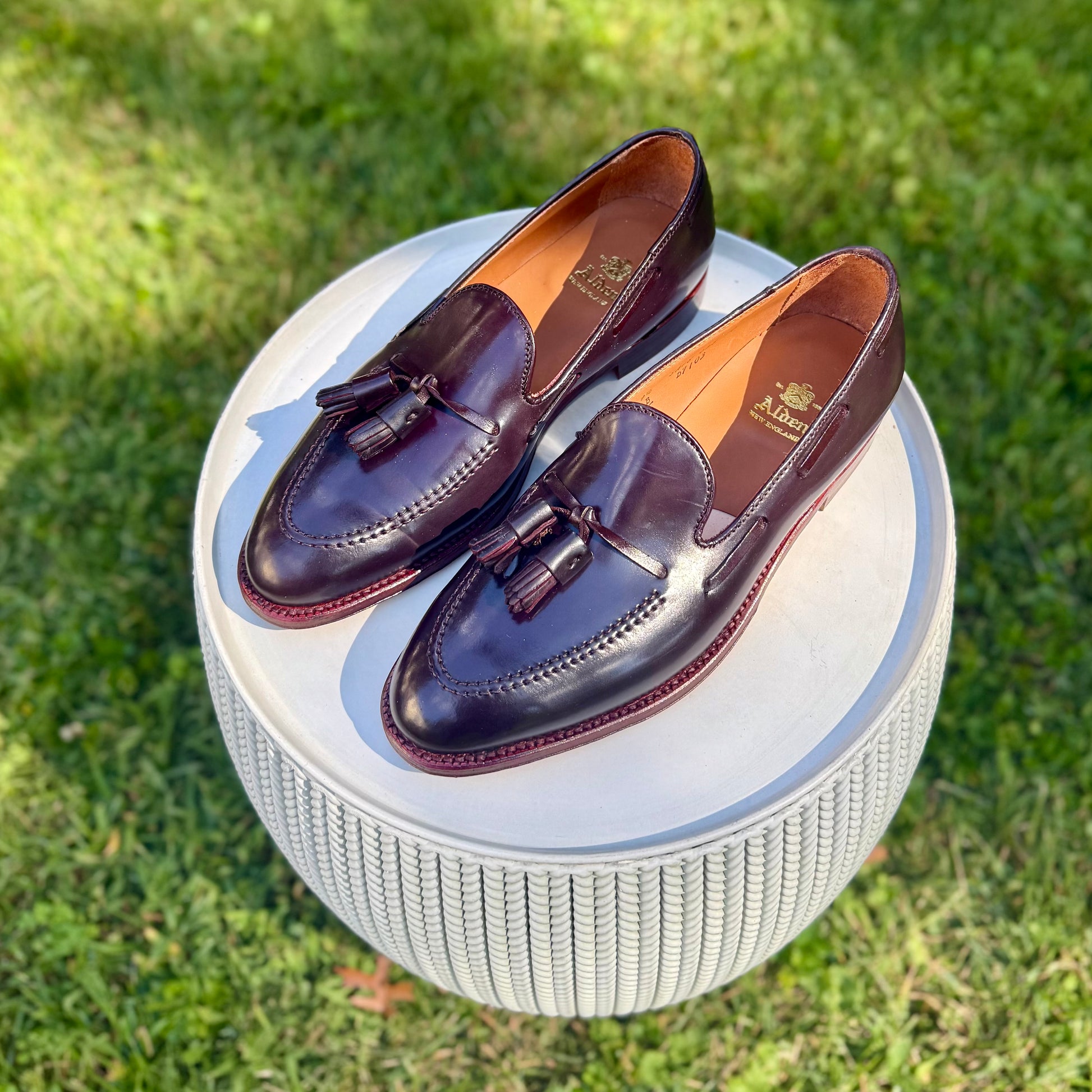 The Alden Madison D7103 - Antique Tassel Loafer in Color 8 Shell Cordovan is displayed on a round, textured white stool outdoors on green grass.