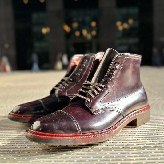 A pair of Alden Madisons D2824HC Cap Toe Boots, crafted from Color 8 Shell with intricate stitching and striking red soles, rest against a textured surface. Their sleek design stands out against a blurred urban backdrop adorned with glimmering lights.