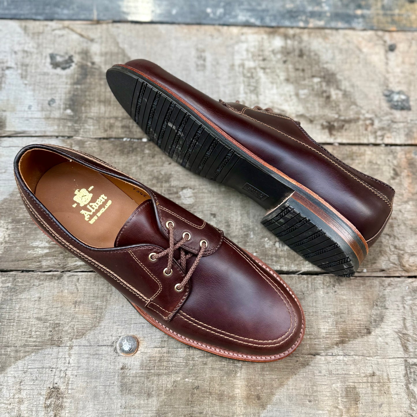 The Alden Madison 734L - Ivy Blucher in Brown Chromexcel, featuring contrast stitching and a Leisure Rubber Sole, is displayed on weathered wood. One shoes interior displays the gold Alden logo.