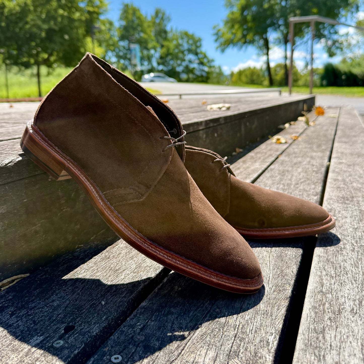 Alden Madison’s 1492 - Unlined Chukka in Brown Suede, featuring a Single Flex Leather Sole and classic Leydon Last, sits outdoors on a wooden deck with trees and blue sky in the background on a sunny day.