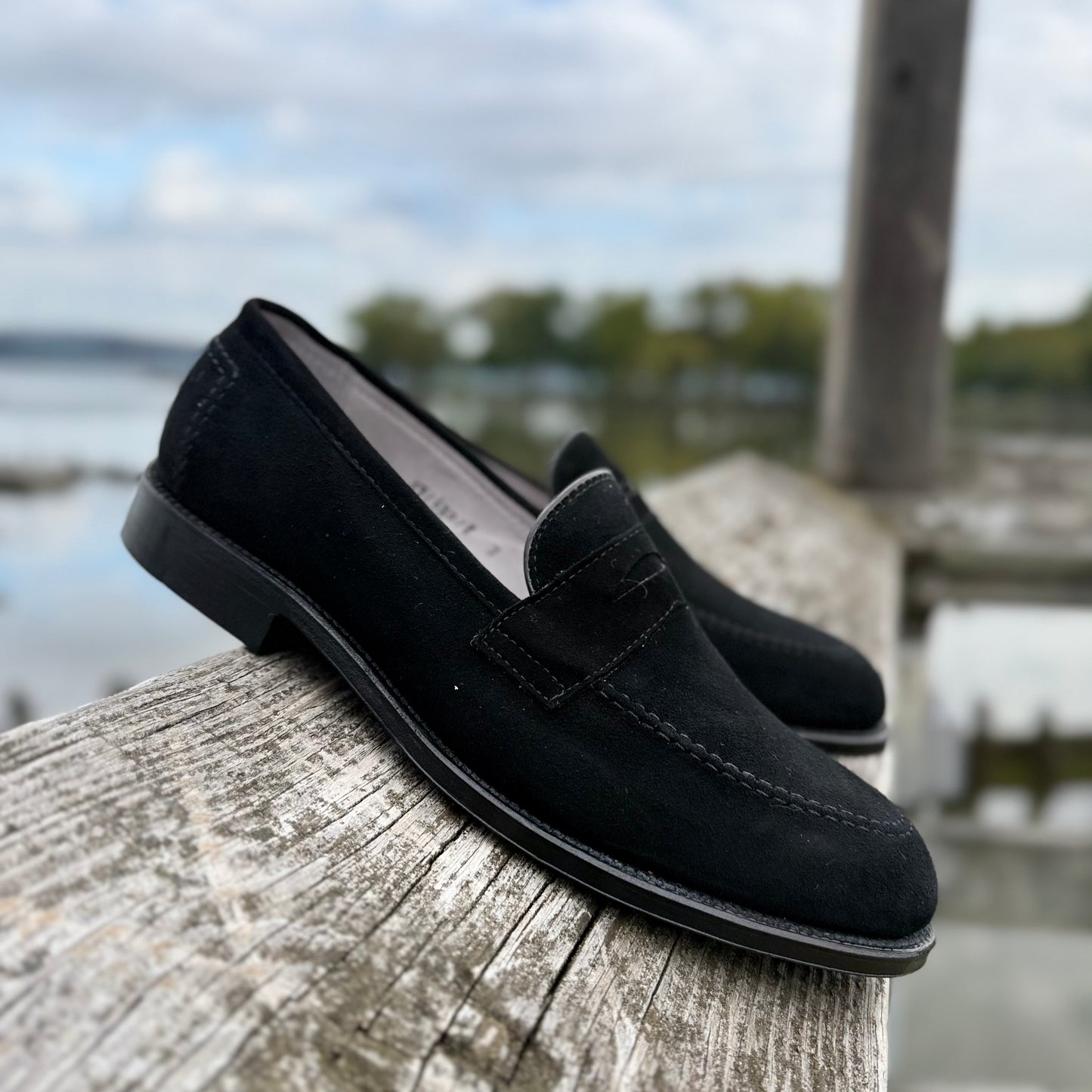 The Alden Madison D2229 “Stuyvesant” Madison Penny in black suede, featuring leather soles, rests on a weathered wooden railing with a blurred outdoor scene of water, trees, and cloudy sky in the background.