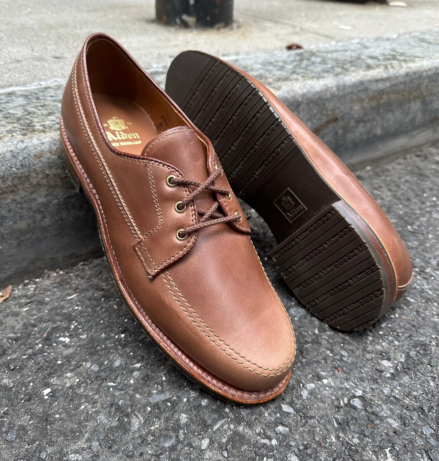 A pair of Alden Madison D5428L - Ivy Blucher shoes in Natural Chromexcel are displayed on a sidewalk, with one shoe standing upright and the other turned to highlight its distinctive Leisure Rubber Sole.