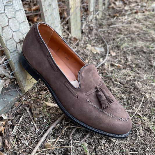 The Alden Madison 666 Tassel Loafer in Brown Suede sits by a wooden fence on grass and dry leaves. Crafted on the Aberdeen Last, it boasts a black leather sole and intricate edge stitching, exuding timeless elegance.