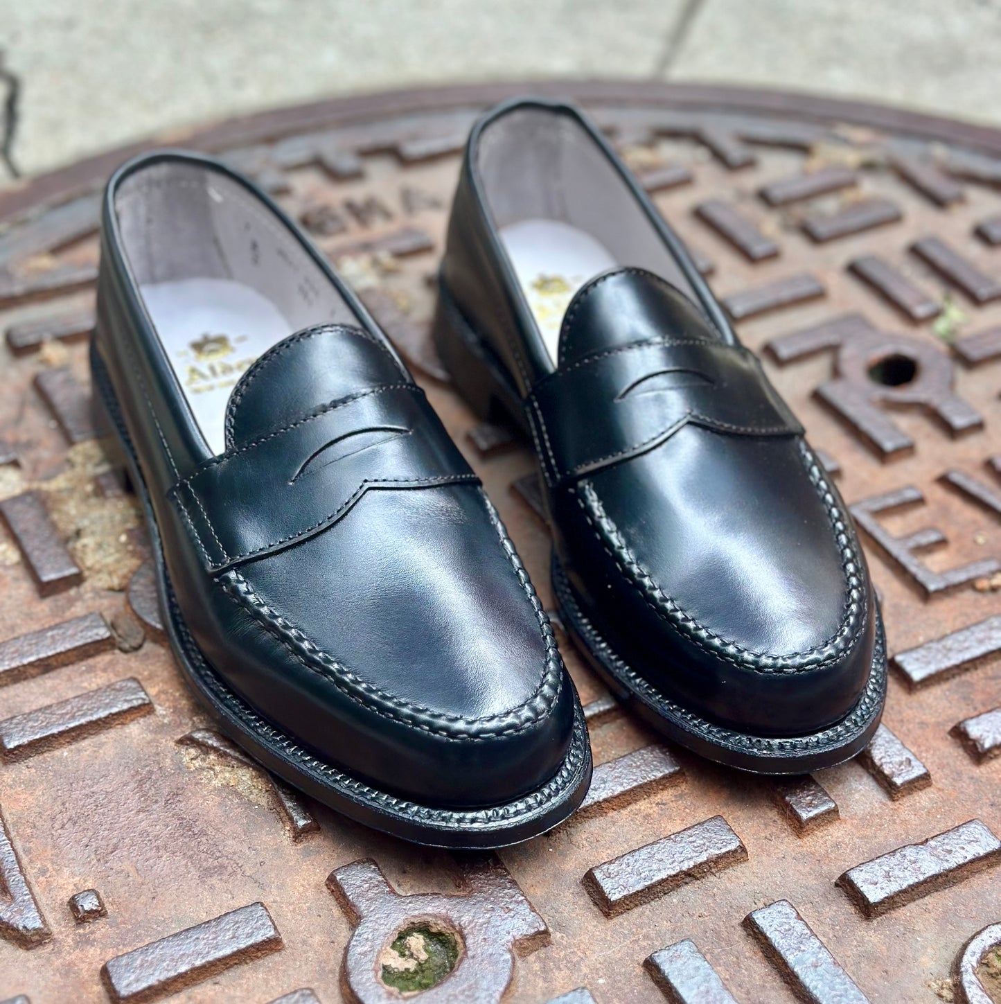 The Alden Madison 981 - Leisure Hand Sewn in Black Calfskin loafers with a Single Oak Leather Outsole are displayed on a textured, round metal manhole cover outdoors.