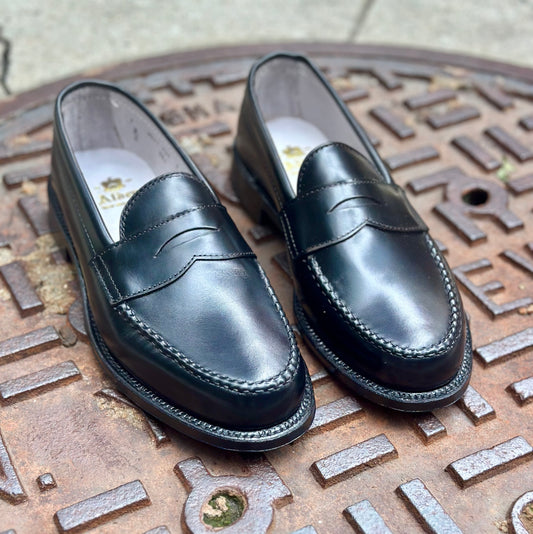 The Alden Madison 981 - Leisure Hand Sewn in Black Calfskin loafers with a Single Oak Leather Outsole are displayed on a textured, round metal manhole cover outdoors.