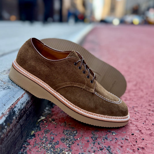 The ALDEN SHOES M7601Y Snuff Suede Indy Shoe with Wedge features tan rubber soles and is elegantly displayed on a textured curb, angled perfectly. In the background, a bustling urban street scene with buildings and traffic is harmoniously blurred.