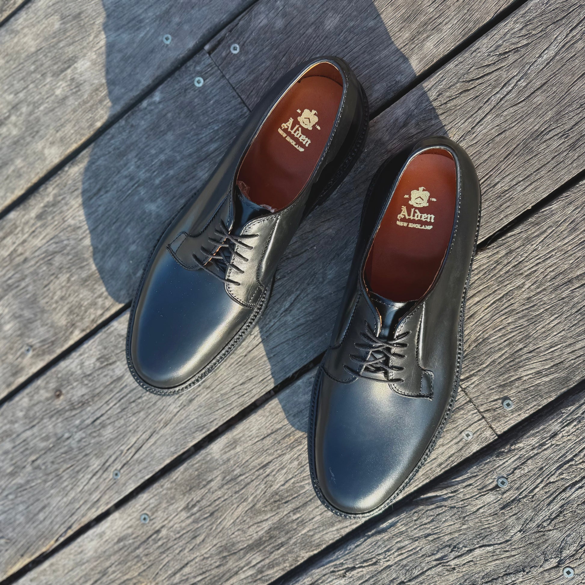 A pair of ALDEN SHOES 9909 Black Calfskin Plain Toe Bluchers, featuring tan insoles with gold logos, displayed on a weathered wooden deck.