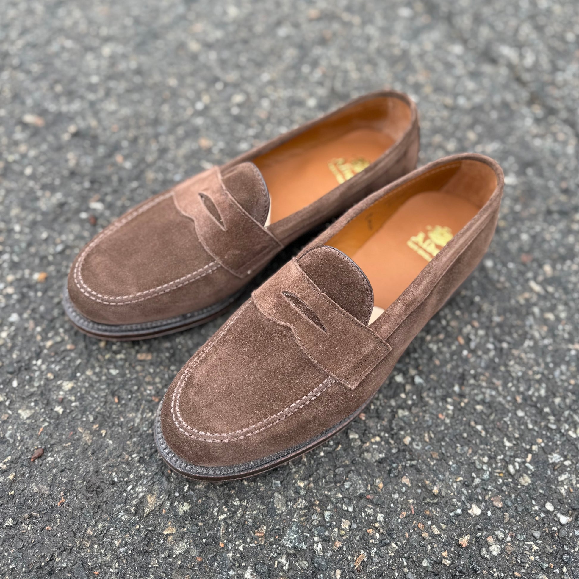 The Alden Madison 6175F - Penny Loafer in Humus Suede, featuring Flex Oiled Leather Soles, is showcased on a rugged asphalt surface.