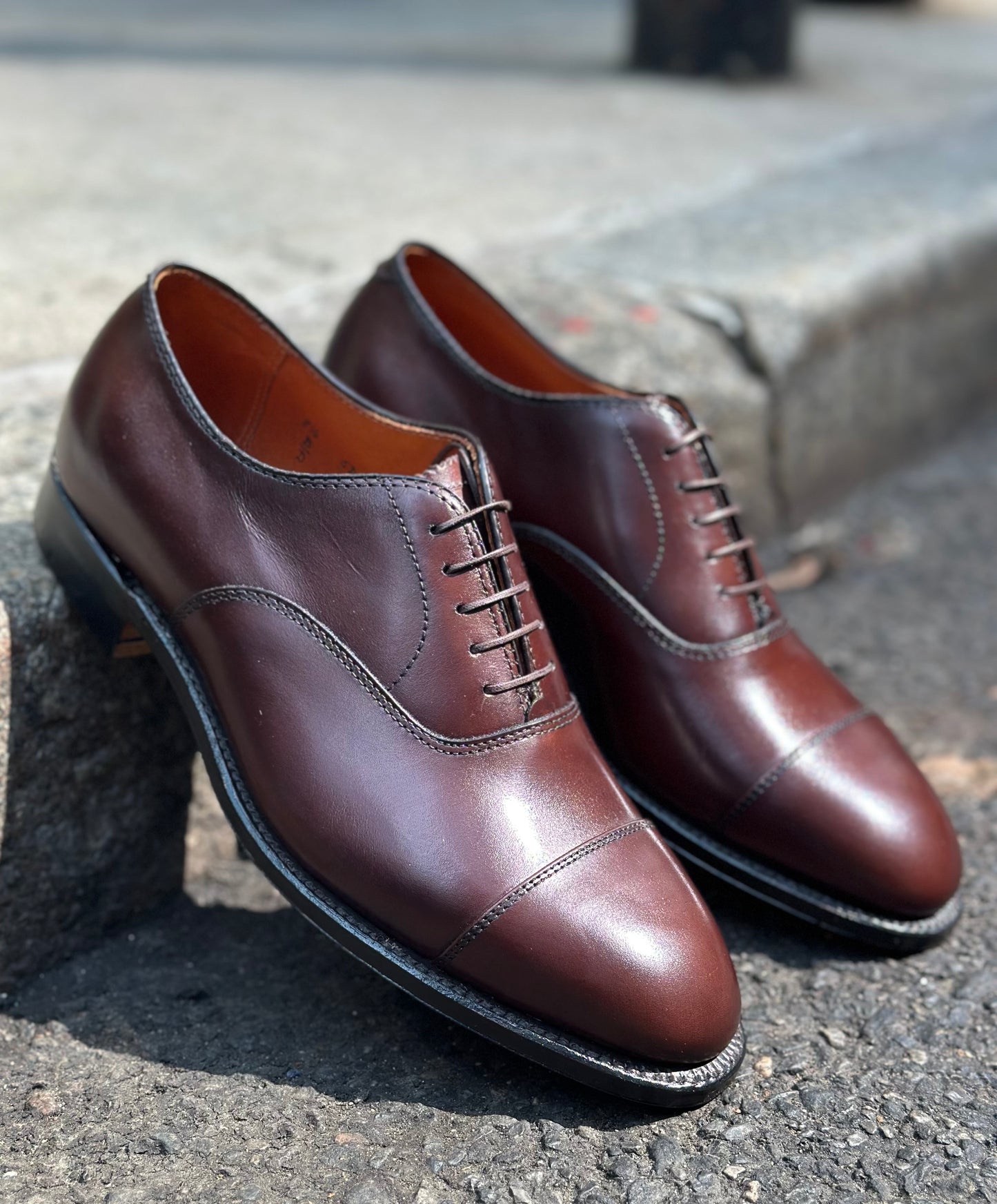 The Alden Madison 920 - Cap Toe Bal in Brown Calfskin, a pair of polished oxford dress shoes with cap toes and leather soles, is shown outdoors on concrete and asphalt.