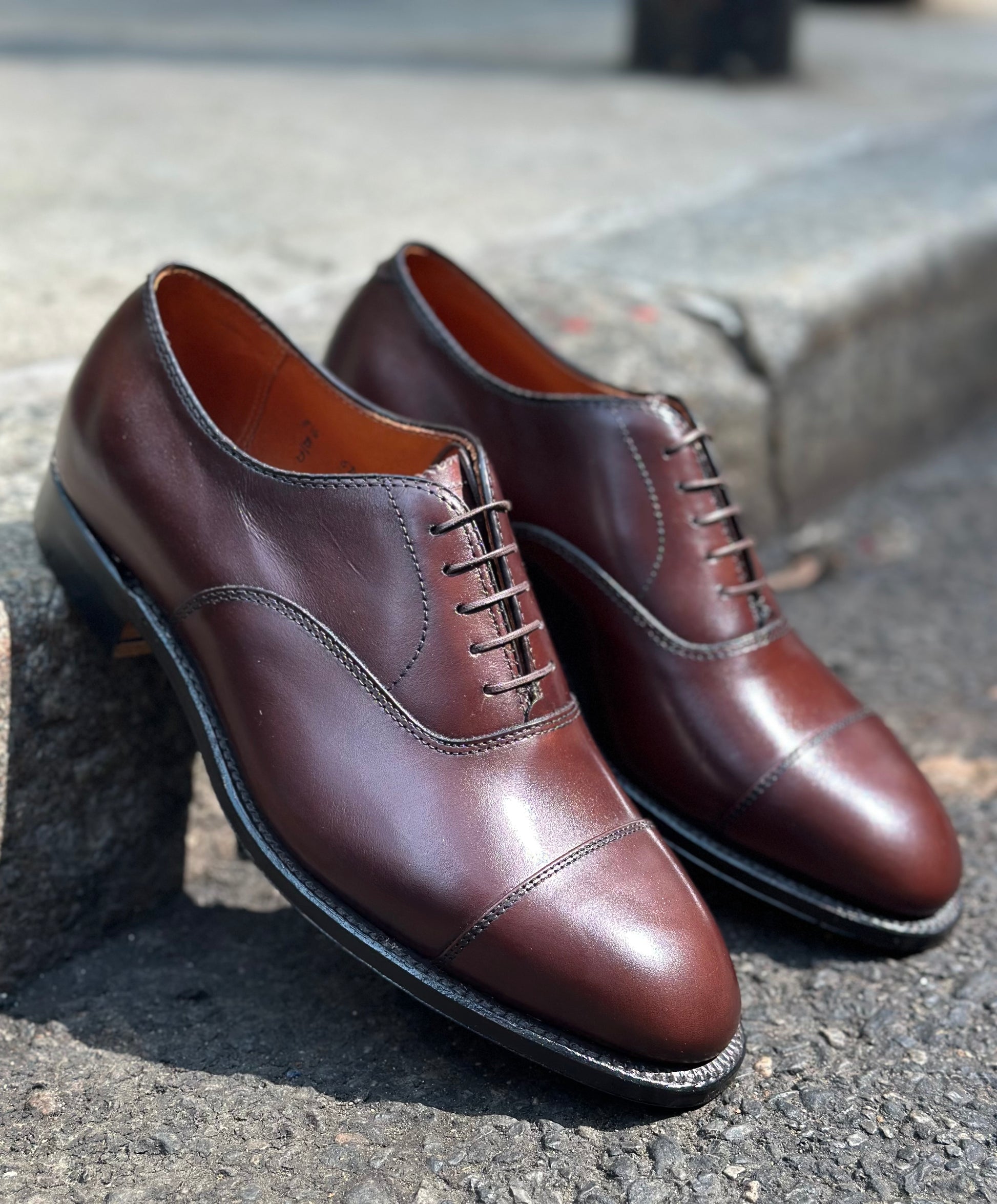 The Alden Madison 920 - Cap Toe Bal in Brown Calfskin, a pair of polished oxford dress shoes with cap toes and leather soles, is shown outdoors on concrete and asphalt.