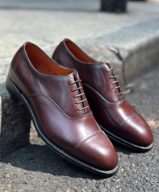 The Alden Madison 920 - Cap Toe Bal in Brown Calfskin, a pair of polished oxford dress shoes with cap toes and leather soles, is shown outdoors on concrete and asphalt.