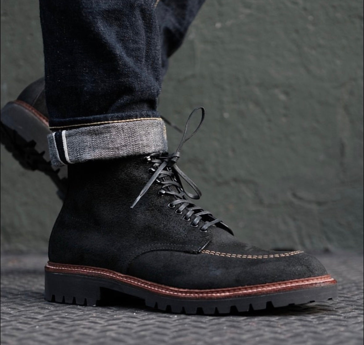 Wearing dark rolled-up jeans and Alden Madisons D9916H - Indy Boot in Earth Reverse Chamois with rugged Vibram Heavy Lug Outsoles and contrast stitching, a person stands on a textured gray surface before a green wall.