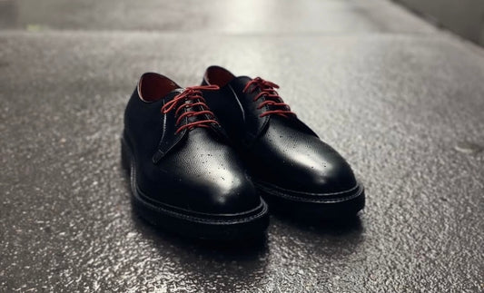 The Alden Madison 949 - All Weather Walker in Black Alpine Grainp dress shoes with red laces rest on wet, textured pavement, water droplets visible on their surface.