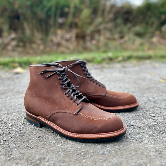 Alden Madisons D2909H Indy Boots feature Tobacco Smooth Chamois leather with precise lacing and a sleek design, complemented by a striking black neocork sole against an outdoor textured surface and a blurred lush green backdrop.