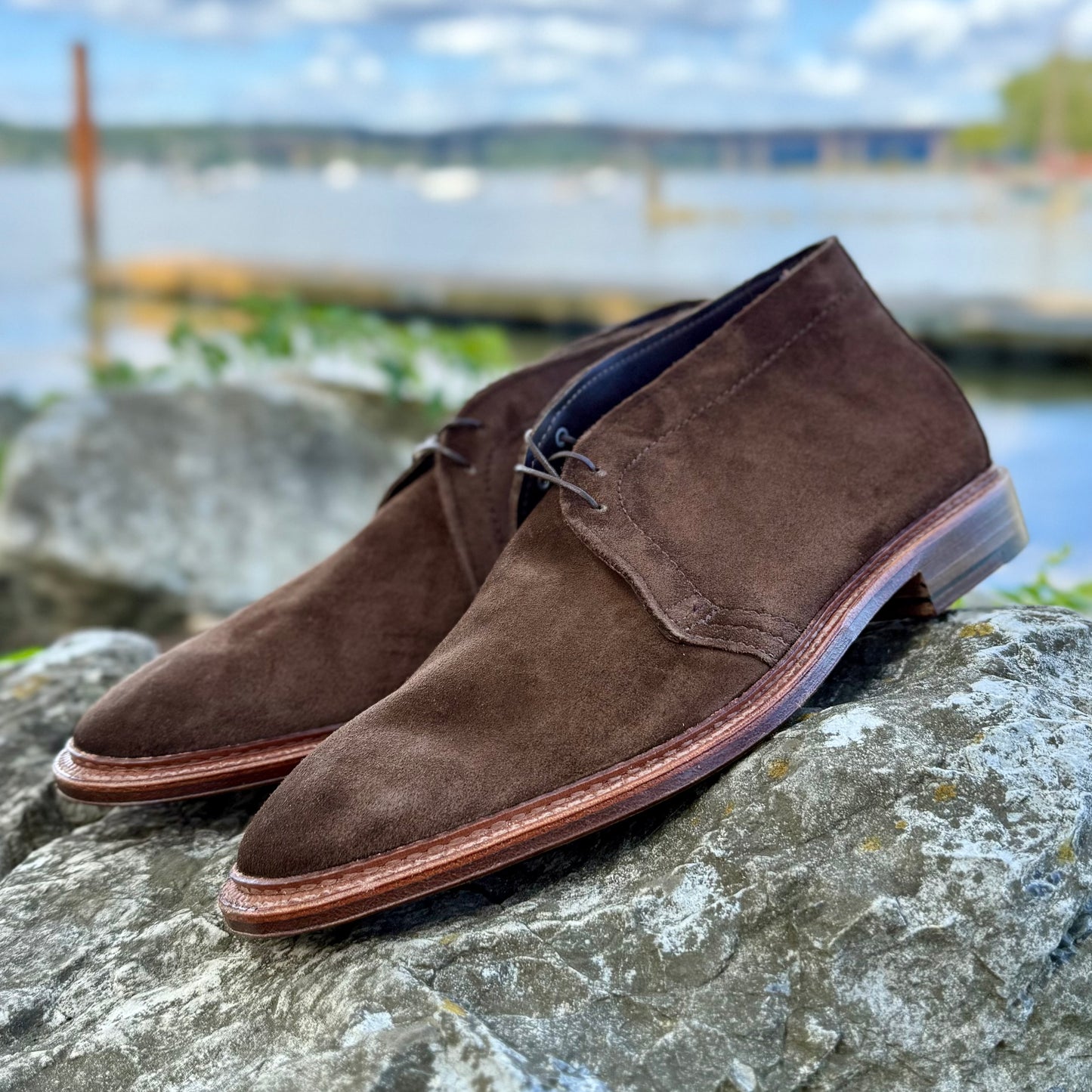 The Alden Madison 1492 - Unlined Chukka in Brown Suede sits on a large gray rock outdoors, with water, trees, and a cloudy sky softly blurred in the background.