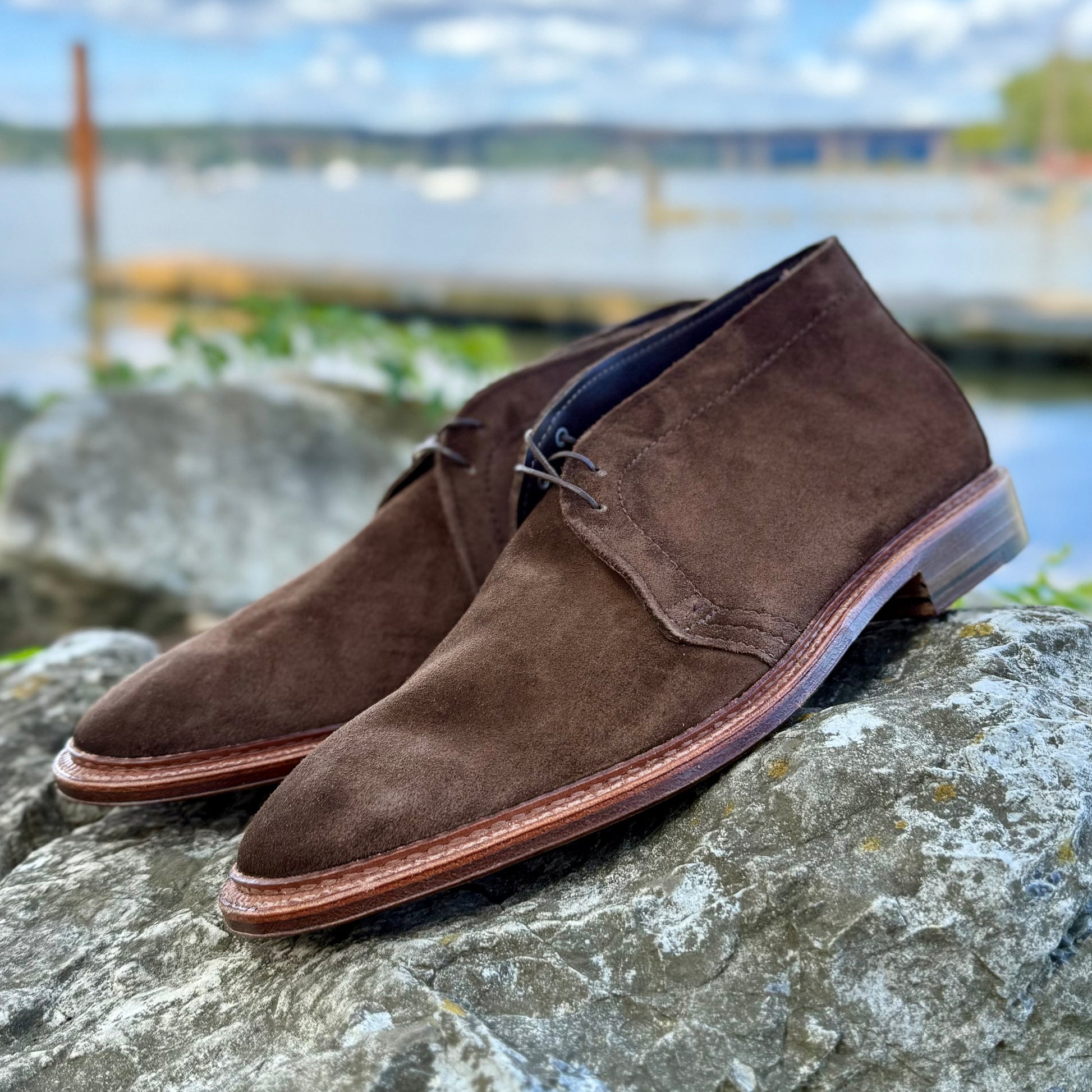 The Alden Madison 1492 - Unlined Chukka in Brown Suede sits on a large gray rock outdoors, with water, trees, and a cloudy sky softly blurred in the background.