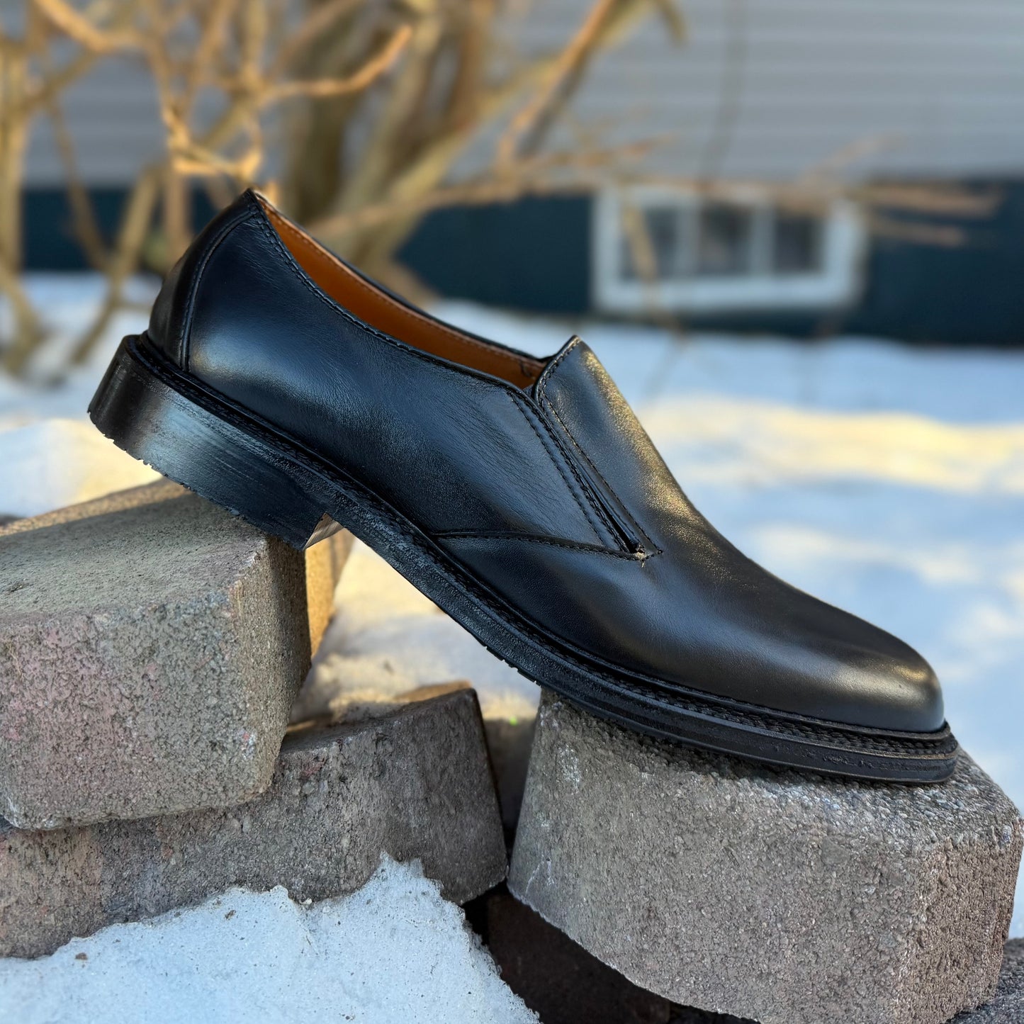 The ALDEN SHOES D2406L "Double Gore" Slip On Rubber Sole, crafted in black calfskin, is shown atop stacked concrete blocks outdoors with snow and a blurred house behind it—a stylish, comfortable choice for any occasion.