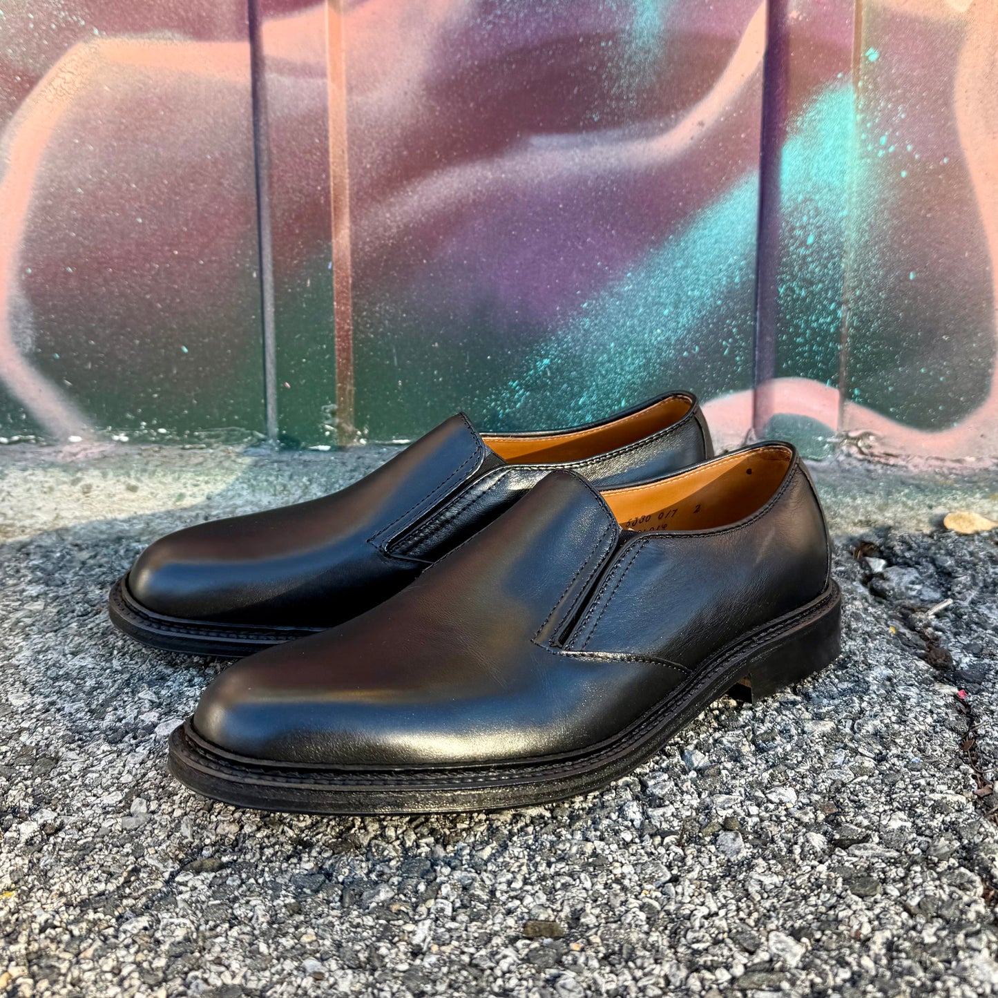 A pair of ALDEN SHOES D2406L "Double Gore" black calfskin slip-on ladies shoes with rubber soles is shown on textured pavement, set against a colorful spray-painted wall.