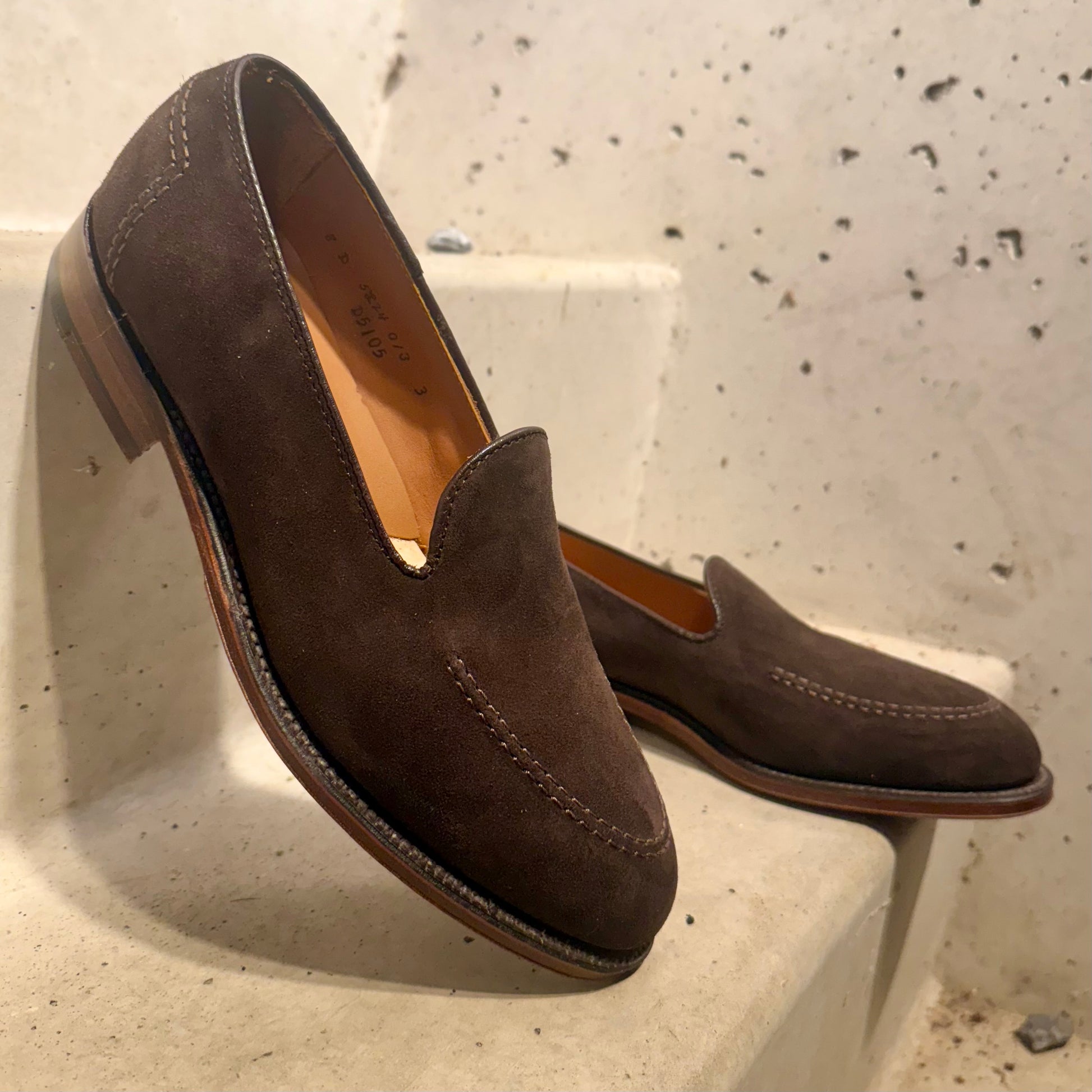 A pair of ALDEN SHOES D5105 - Vanderbilt in Dark Brown Suede with stitching and a Flex Leather Sole are displayed on beige concrete steps against a lightly speckled wall.