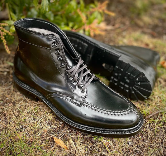 The Alden Madison 40219HC - NST Tanker Boot in Black Shell Cordovan, with visible stitching and a sturdy Commando Sole, is shown on grass—one boot upright, the other lying on its side amid greenery.