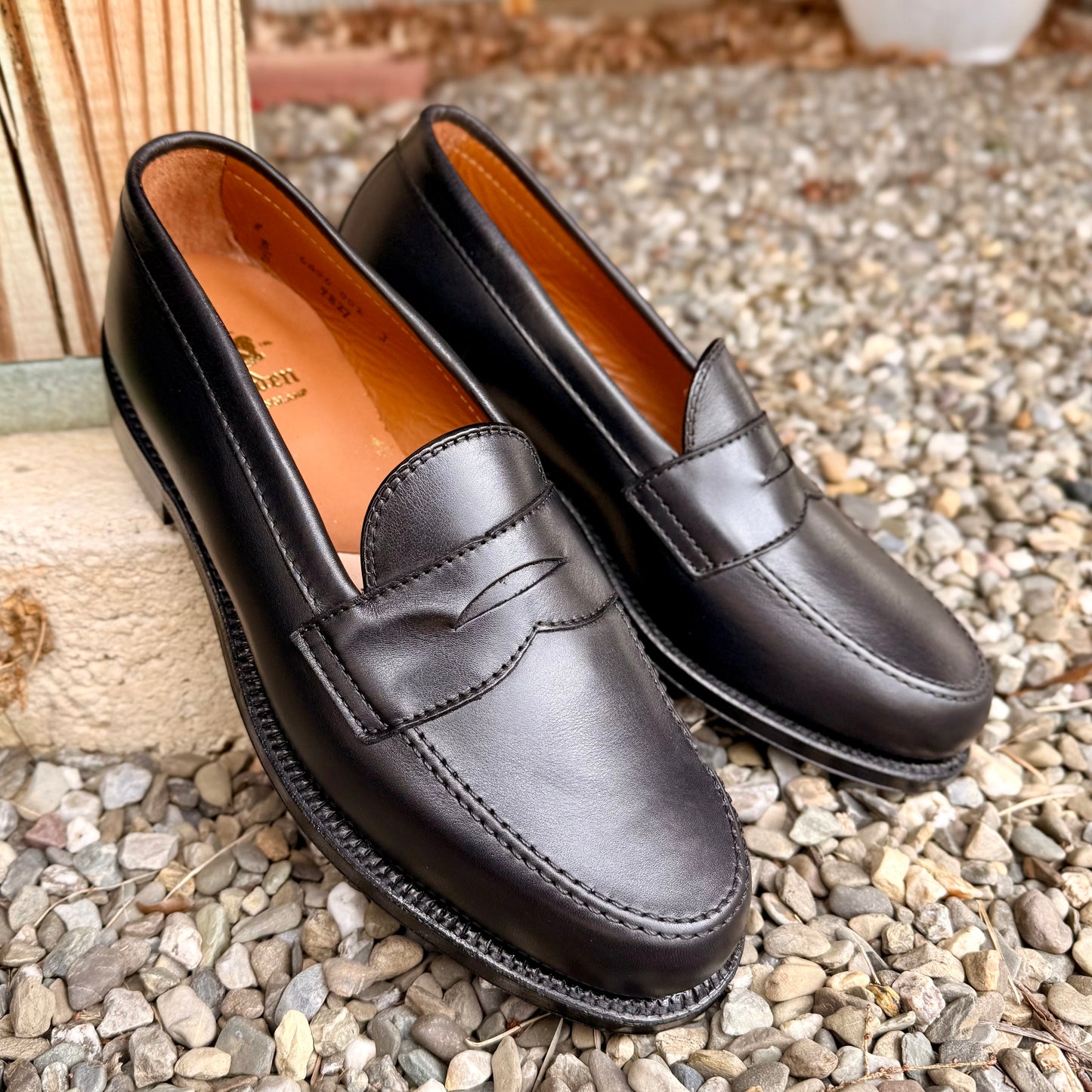 The 9827 - Black Calf Leisure Penny Loafer by ALDEN SHOES, featuring brown insoles and a single leather sole, is displayed on multicolored gravel near a wooden post, highlighting its classic silhouette.