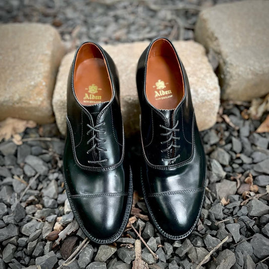 A pair of Alden Madison 9071 - Straight Tip Bal dress shoes in Black Shell Cordovan sit side by side on dark gravel, with two rectangular stone blocks behind them.