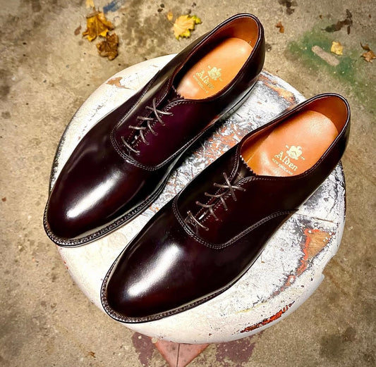 A pair of Alden Madison 934 - Plain Toe Bal in Color 8 Shell Cordovan dress shoes with laces, featuring branded insoles and a refined Single Oak Leather Sole, rests on a round, weathered white stool with colorful paint marks.