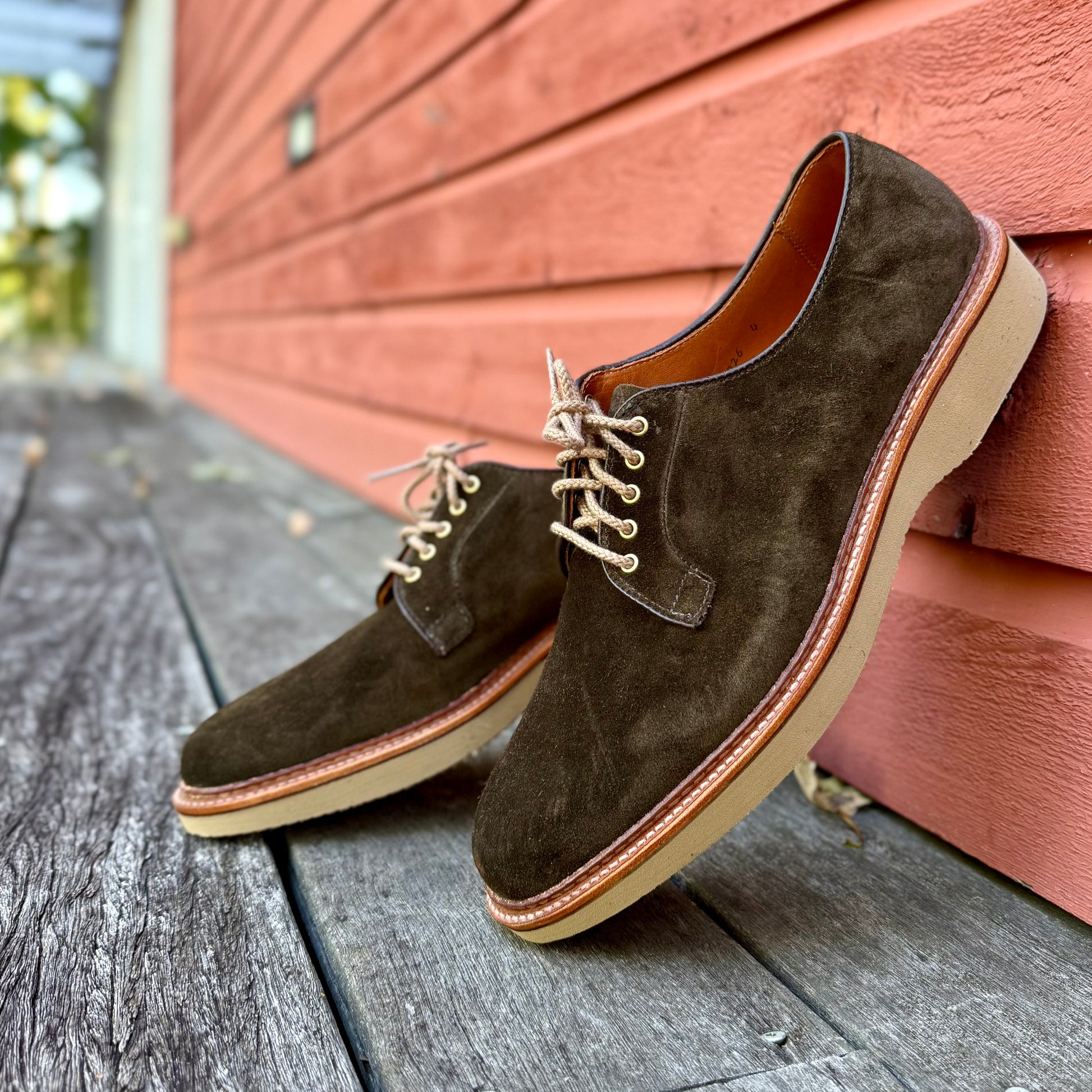 A pair of ALDEN SHOES D5426 - Loden Suede Plain Toe Blucher with a tan wedge sole rests on a weathered wooden surface, propped against a red wooden wall.