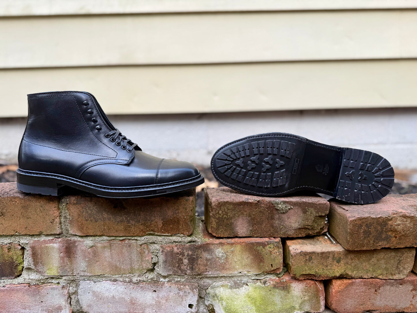 The Alden Madison 40797HC Black Cap Toe Boot with Commando Sole is shown on a weathered brick wall—one boot upright, the other on its side to highlight the rugged sole—with a light yellow house wall in the background.