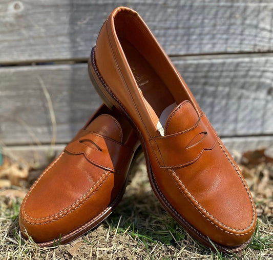 A pair of Alden Madison 983 - Leisure Hand Sewn Burnish Tan Calfskin loafers with leather outsole is displayed outdoors on grass and dry leaves, one shoe upright and the other leaning, with a wooden fence in the background.
