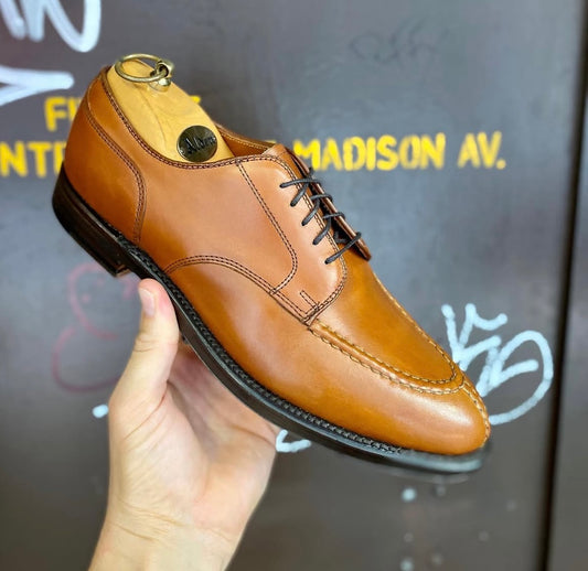 A hand holds up the Alden Madison 962 - Norwegian Split Toe in burnished tan calf leather with black laces and stitching, crafted on the Aberdeen Last, in front of a dark wall with yellow and white graffiti-style text.